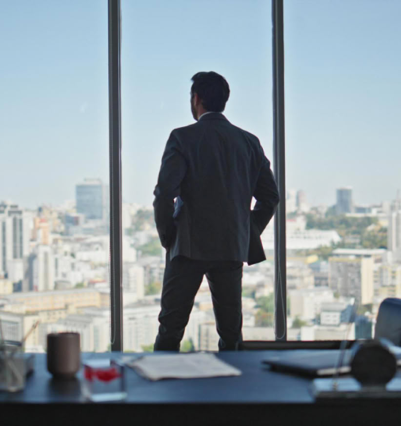 Man in a suit standing in front of large windows overlooking a cityscape.