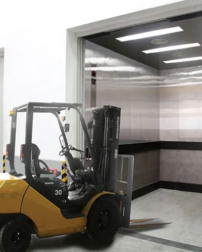 A yellow forklift entering a large freight elevator in a warehouse setting.