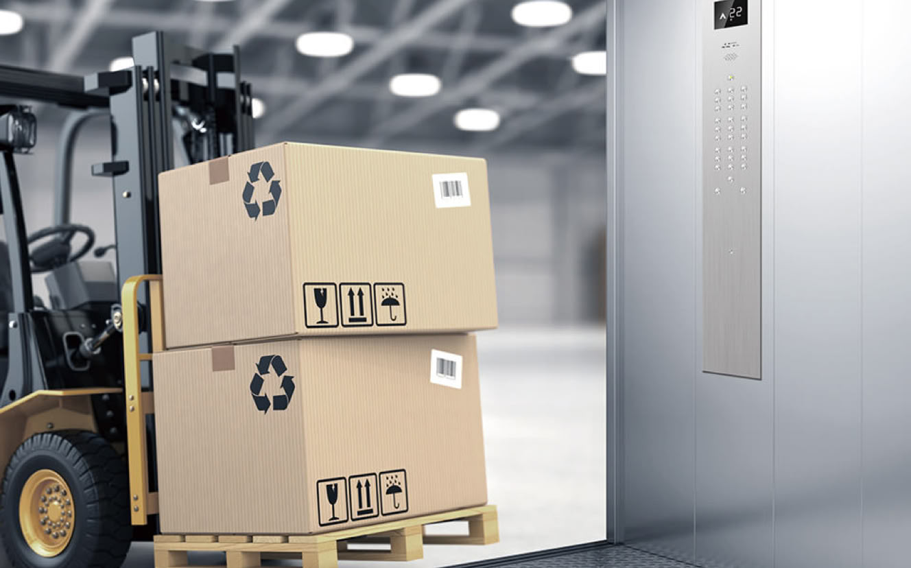 A forklift carrying two stacked cardboard boxes with recycling symbols approaches an elevator in a warehouse.