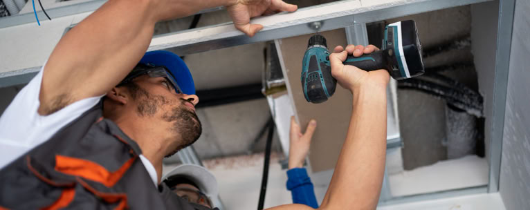 A worker in a blue cap using a power drill to install or repair a ceiling fixture.