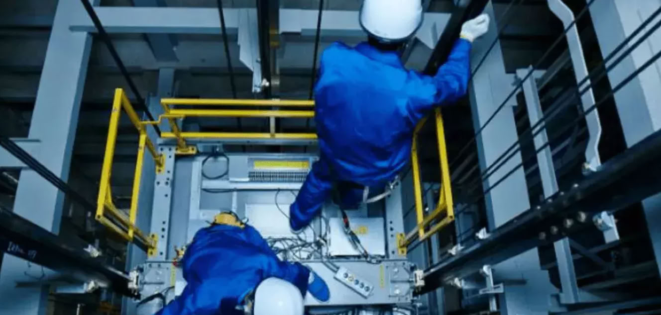 Two workers in blue uniforms and helmets performing maintenance inside an elevator shaft.