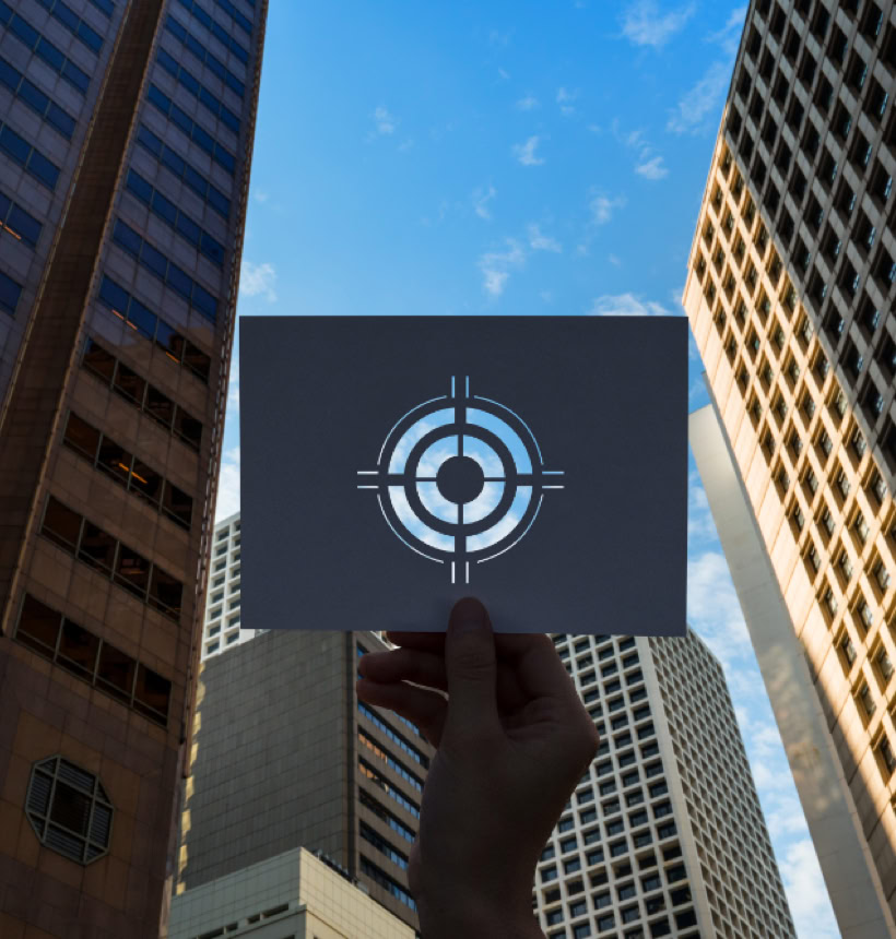 A hand holding a transparent sheet with a target symbol, positioned against a backdrop of tall buildings and a blue sky.